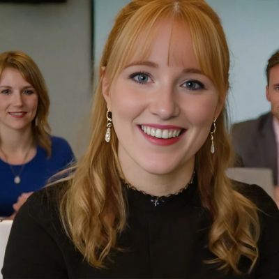 Freundliche junge Frau lächelt in die Kamera, zwei Personen im Hintergrund.