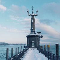 Schneebedeckte Statue am Seeufer mit Mole und gelben Pfählen im Winter.