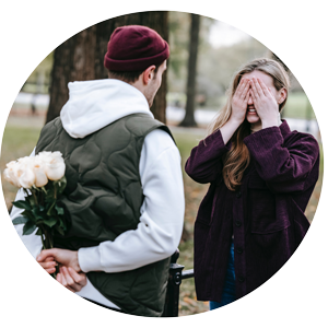 Mann versteckt Blumenstrauß hinter Rücken, Frau hält Hände vor Gesicht im Park.